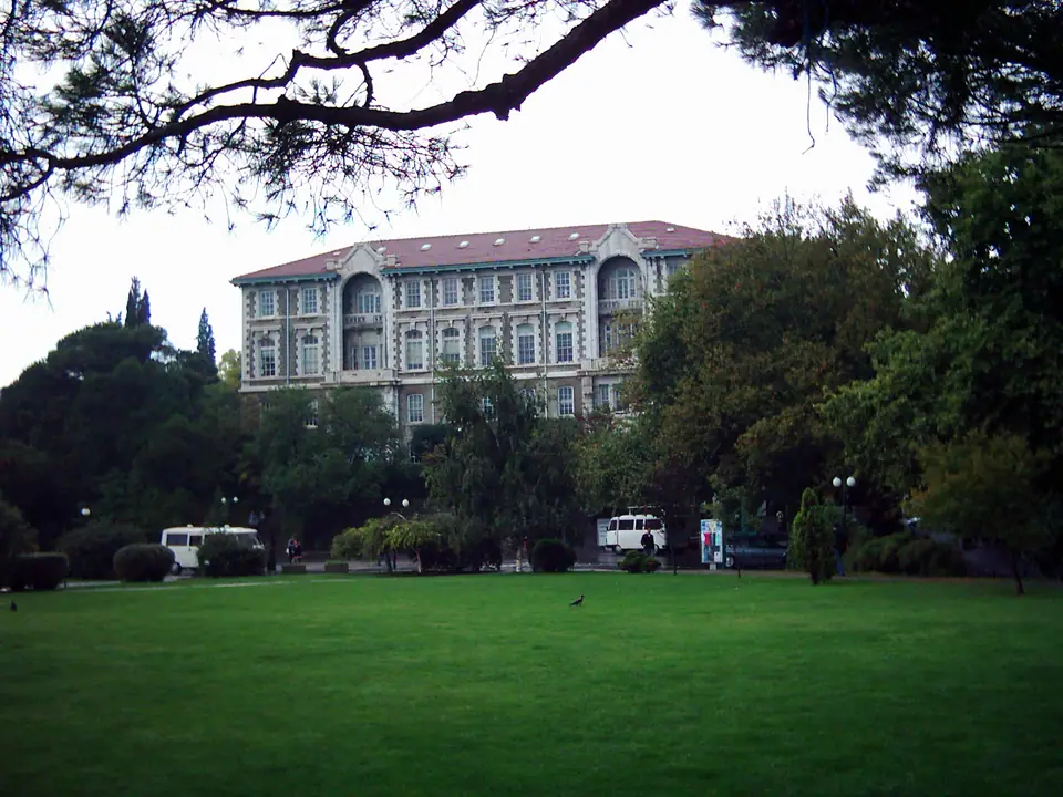 Ağaçlar arasında taş cepheli üniversite binası ve önündeki çimenlik